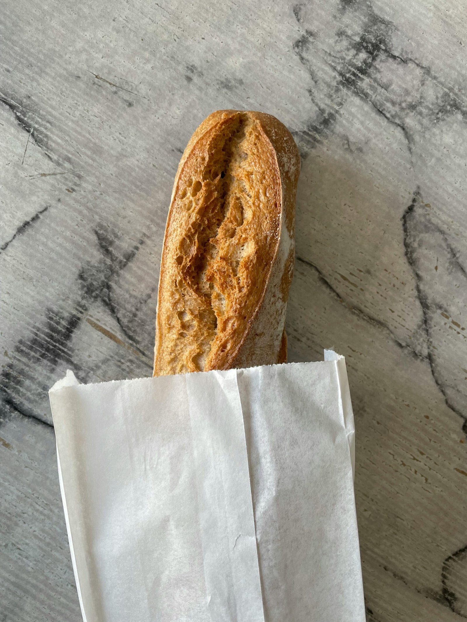 pexels-photo-16652770-16652770 Crispy French baguette in a paper bag on a textured marble surface.