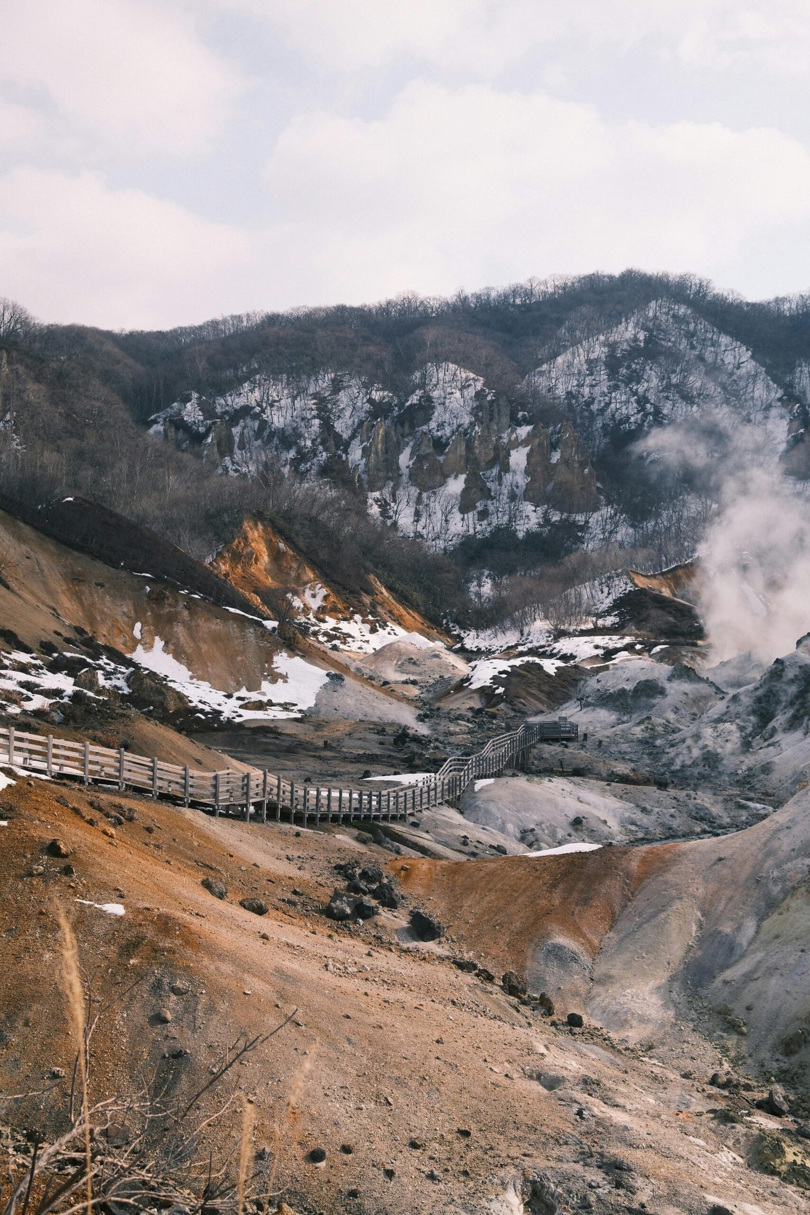 Scenic winter view of Hokkaido's geothermal landscape with wooden walkway and steam rising.