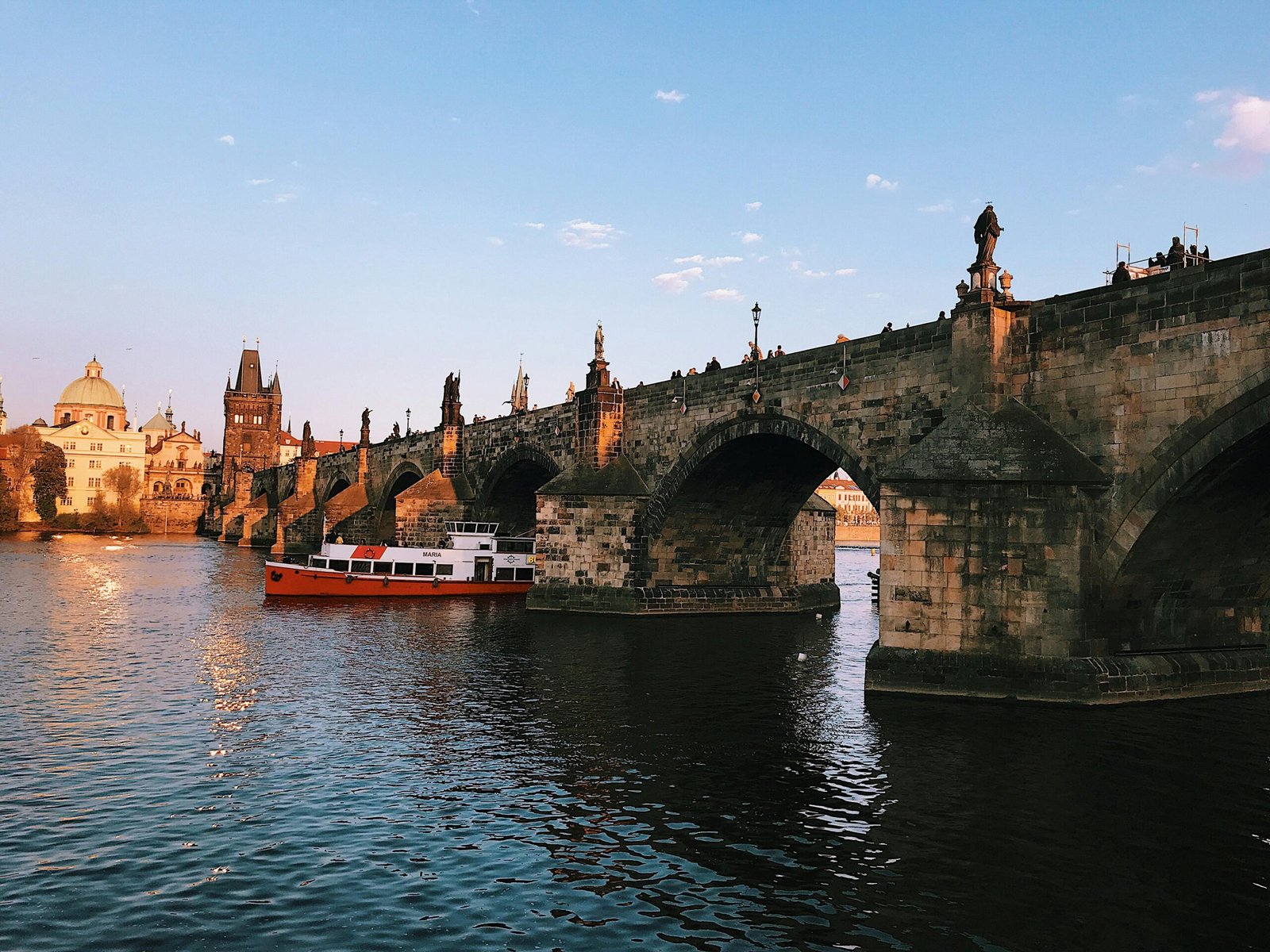 Scenic view of the Charles Bridge and iconic Prague landmarks during sunset with a boat on the Vltava River.